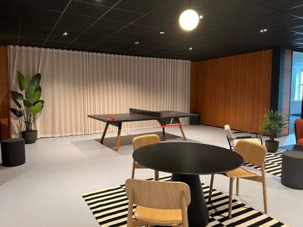 Moderne ontspanningsruimte met een tafeltennistafel, ronde tafel met stoelen, planten en een zwart-wit gestreept vloerkleed.